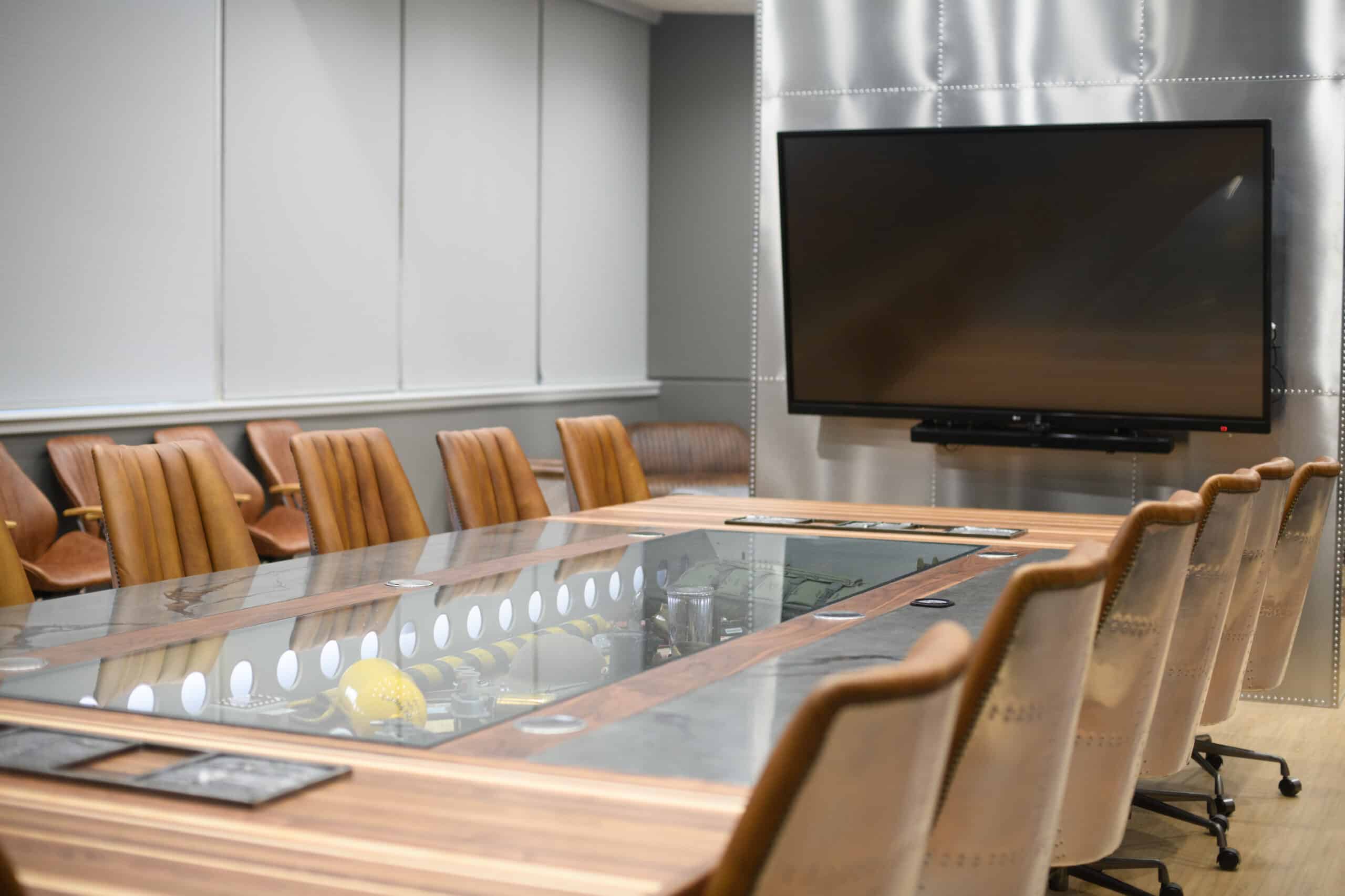 An empty boardroom with a glass-top table, camel-colored chairs, and a large digital screen.