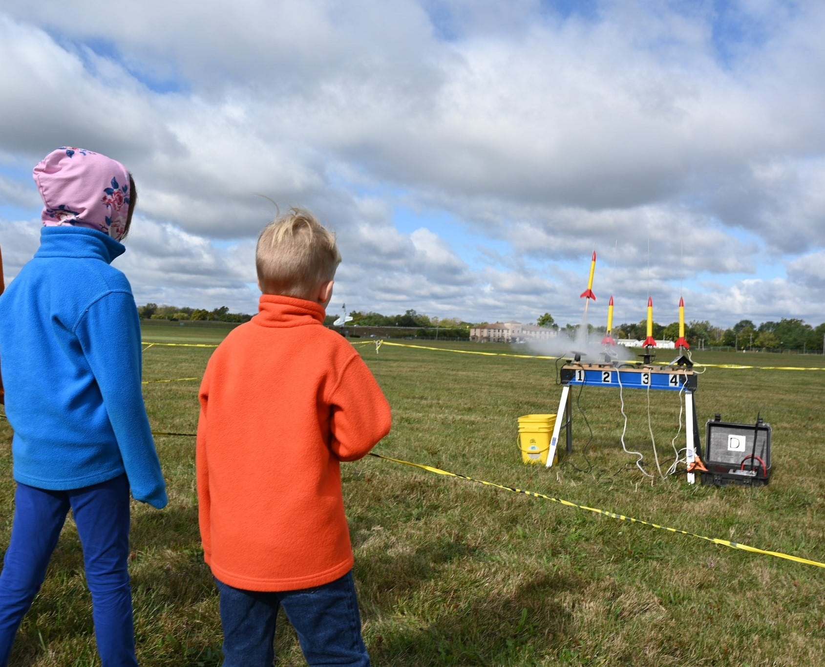 Two children in jackets watching an outdoor model rocket launch.